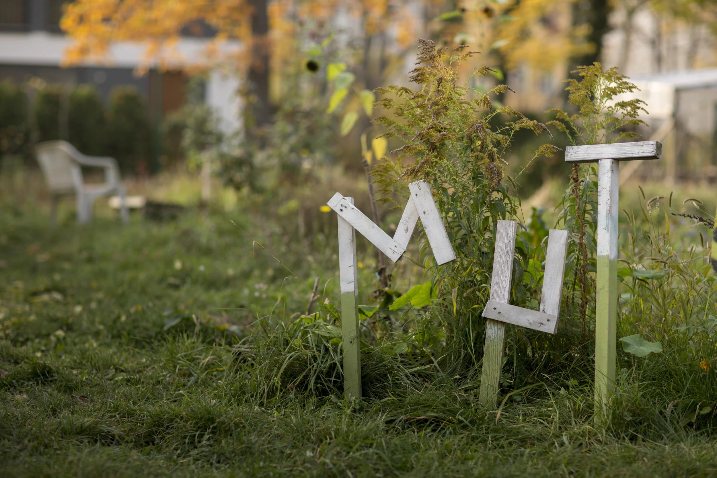 Weiße Holzbuchstaben, die das Wort 'MUT' im Gras der Nachbarschaftsgärten formen, mit einer Gartenbank im unscharfen Hintergrund und herbstlicher Vegetation rundherum.