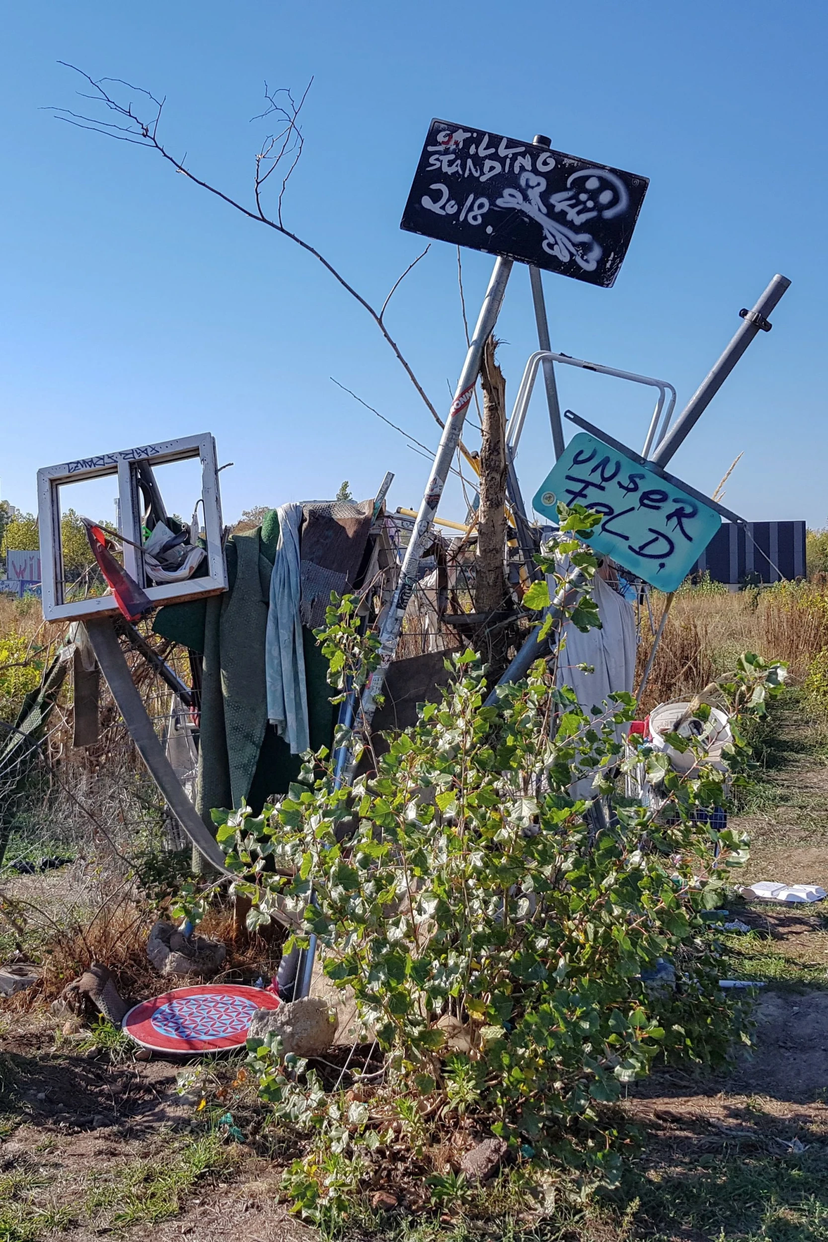 Eine Outdoor-Installation aus diversen Objekten und Schildern mit Schriftzügen 'Still Standing 2018' und 'UNSER FELD' auf dem Jahrtausendfeld in Leipzig Lindenau unter klarem Himmel.