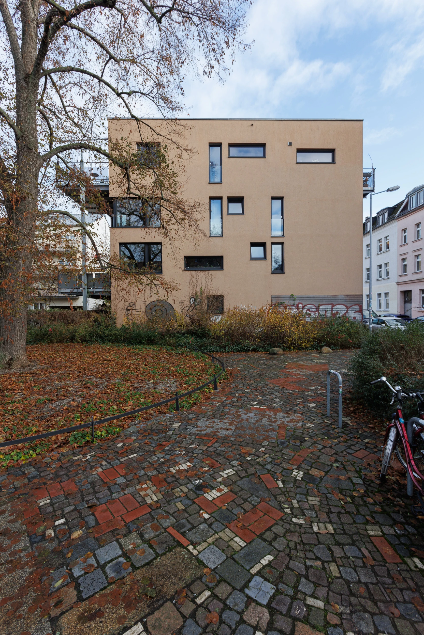 Ein gepflasterter Weg mit Herbstlaub im Vordergrund, der zu einem modernen, beigen Gebäude am Westentaschenpark in Leipzig Lindenau führt, mit einem geparkten Fahrrad an der Seite und Bäumen im Hintergrund.